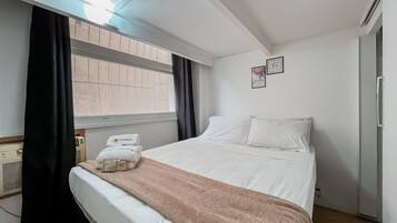 A hotel room with a bed, a window with curtains, a radiator, and a wall with two framed pictures.