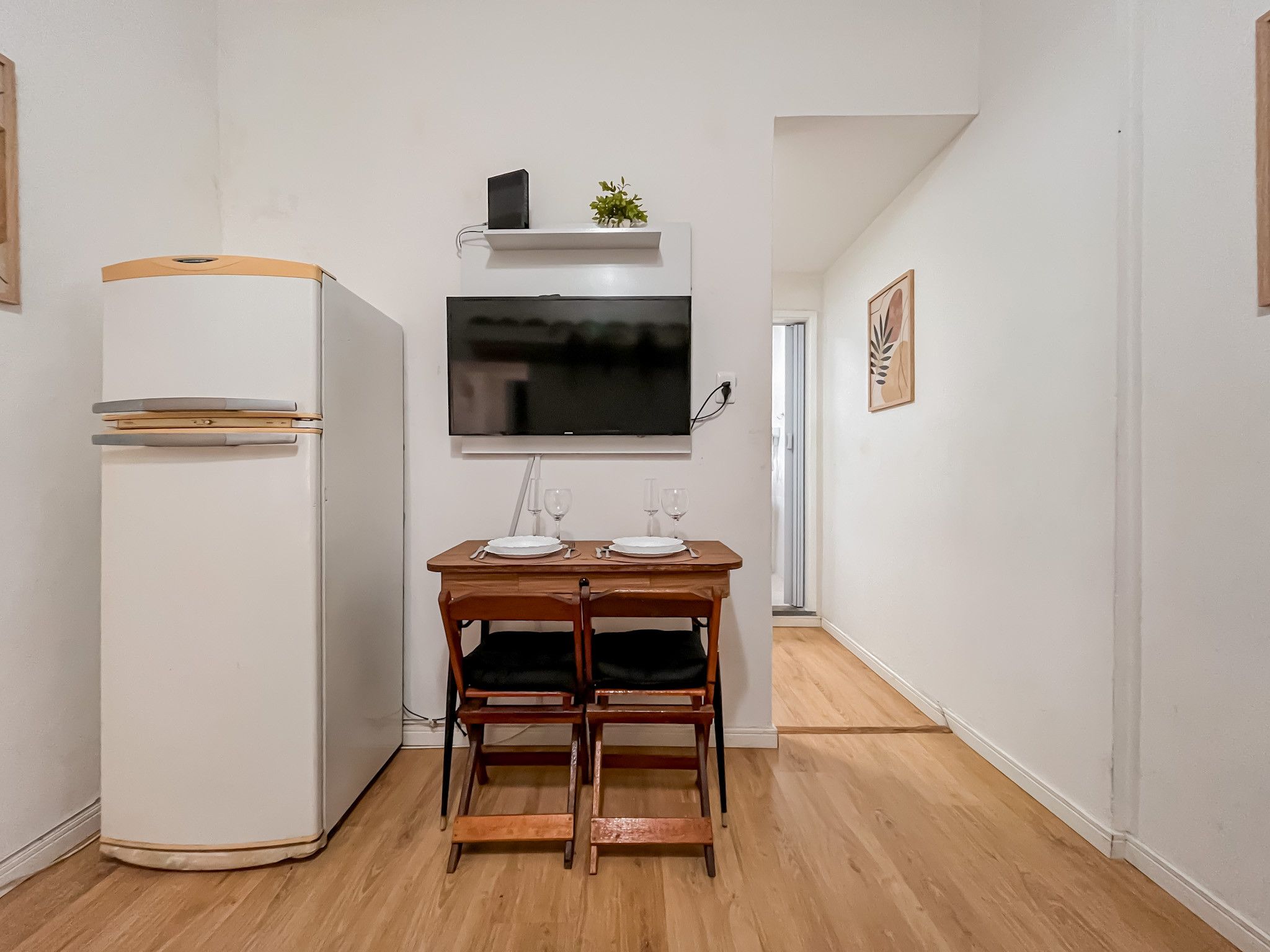 A compact kitchenette with a white refrigerator, a flat-screen TV mounted above a wooden table set for two, and a small potted plant on the counter.