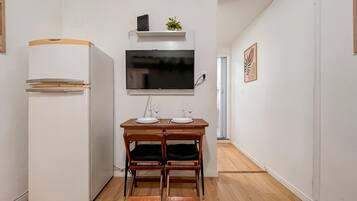 A compact kitchenette with a white refrigerator, a flat-screen TV mounted above a wooden table set for two, and a small potted plant on the counter.