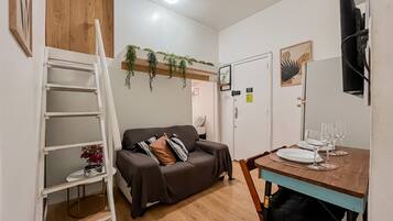 A compact living space with a sofa, a dining table set for two, a ladder, and a small shelf with a plant.