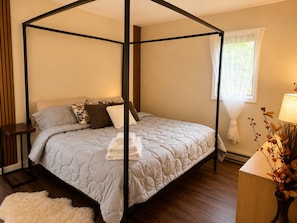 A four-poster bed with a grey quilt, a wooden nightstand, a white fluffy rug, and a bedside table with a lamp and decorative plant. - Spacious 4BR Pocono Chalet - Hot Tub, Barrel Sauna & Game Room (Pocono Summit)