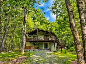Una cabana de dues plantes amb un balcó, envoltada d'arbres i un camí de grava.