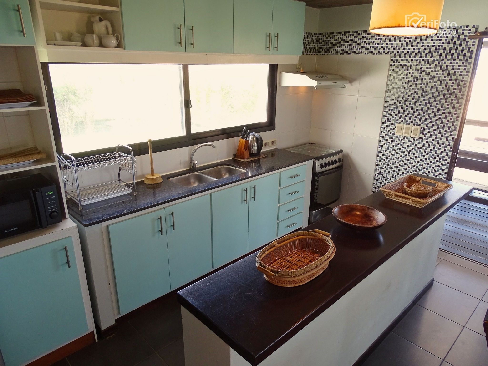 Een keuken met blauwgroene kastjes, een zwart aanrecht, een magnetron, een fornuis en een raam met uitzicht op bomen.