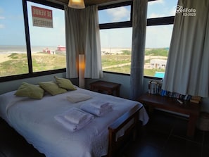A bedroom with a large bed, pillows, a bedside table with books, a lamp, and a view of the landscape.