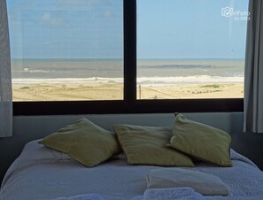 A bed with pillows and a view of the beach through a window.