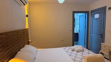 Habitación de hotel con cama, mesita de noche, equipo de aire acondicionado, lámpara colgante y puerta que da a otra habitación.