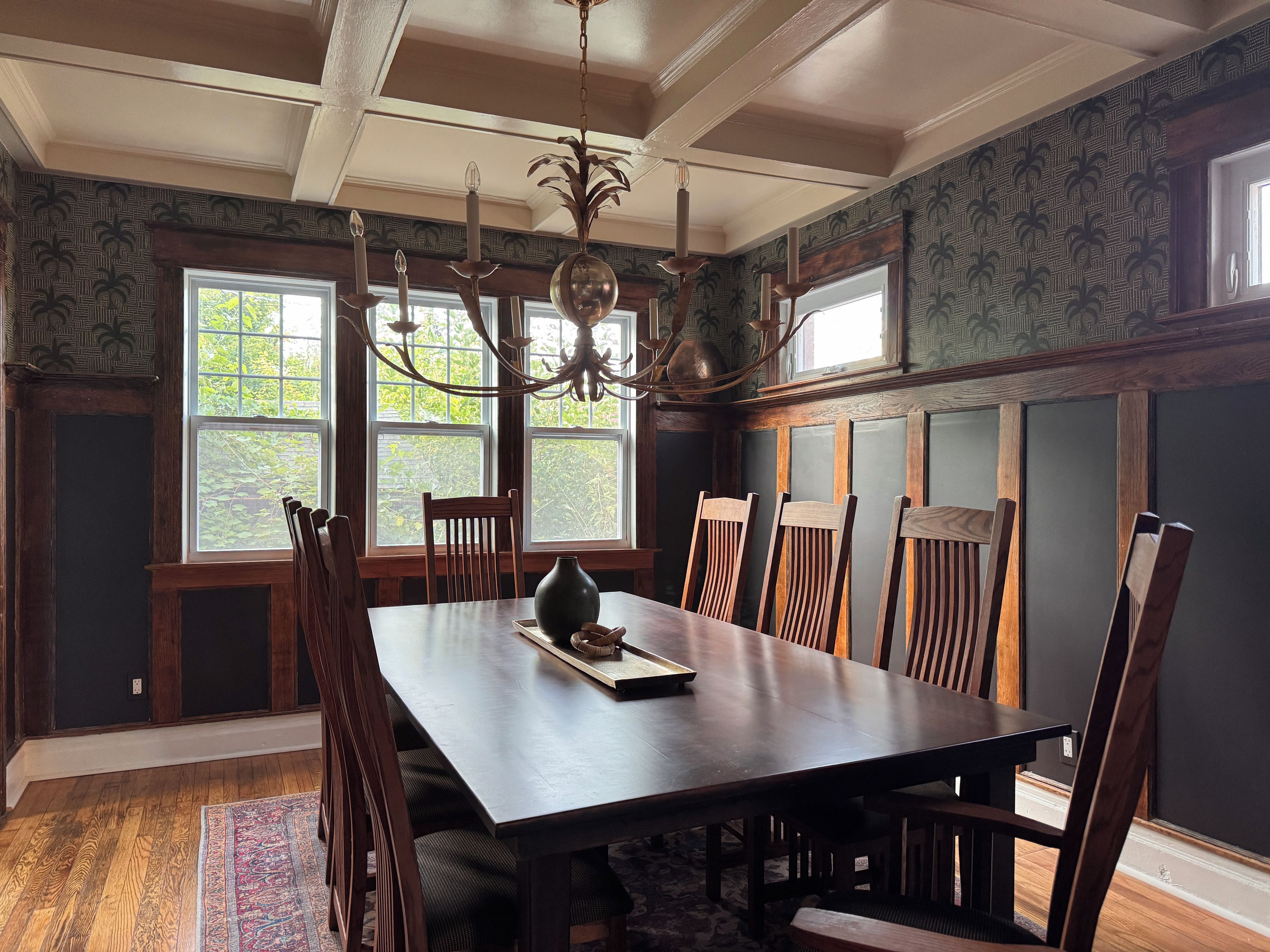 Een eetkamer met een grote houten tafel, meerdere stoelen, een kroonluchter en ramen met uitzicht op groen.