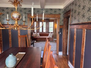 A dining area with a wooden table, chairs, and a vase. There is a sofa and a window in the background.