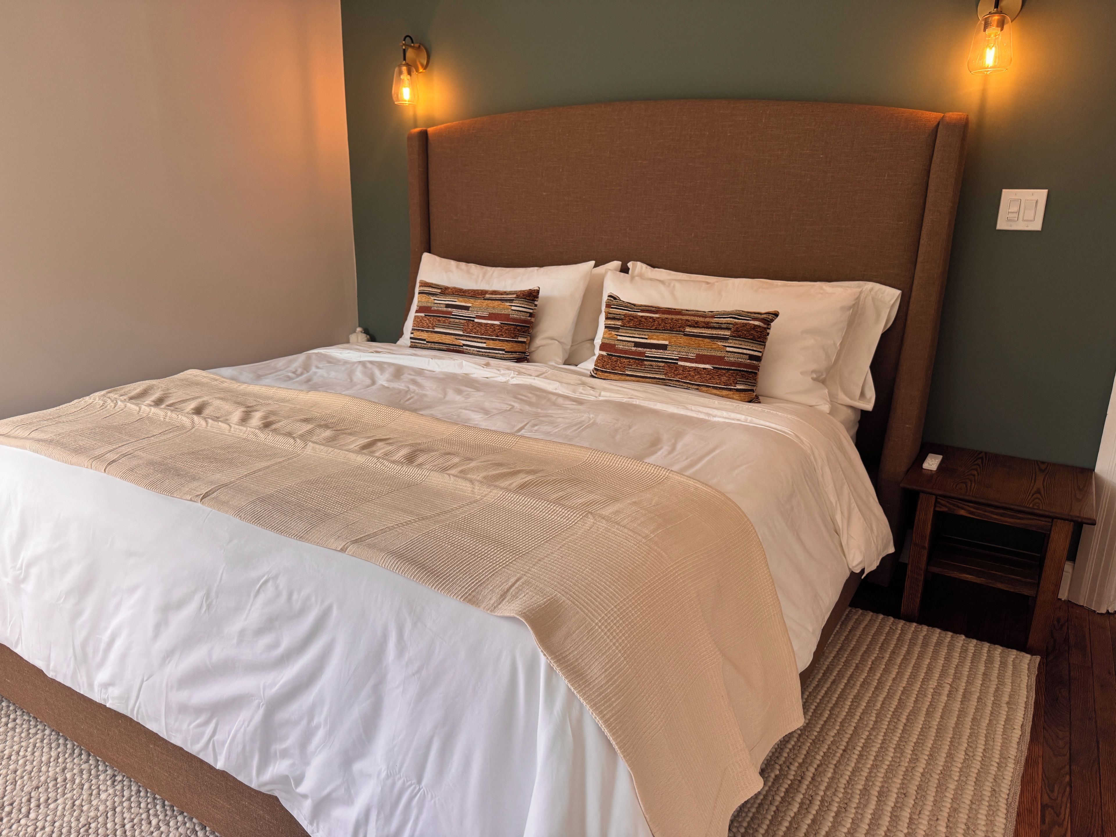Een net opgemaakt bed met een beige dekbedovertrek en twee gestreepte kussens, een houten hoofdbord en een klein nachtkastje met een lamp.