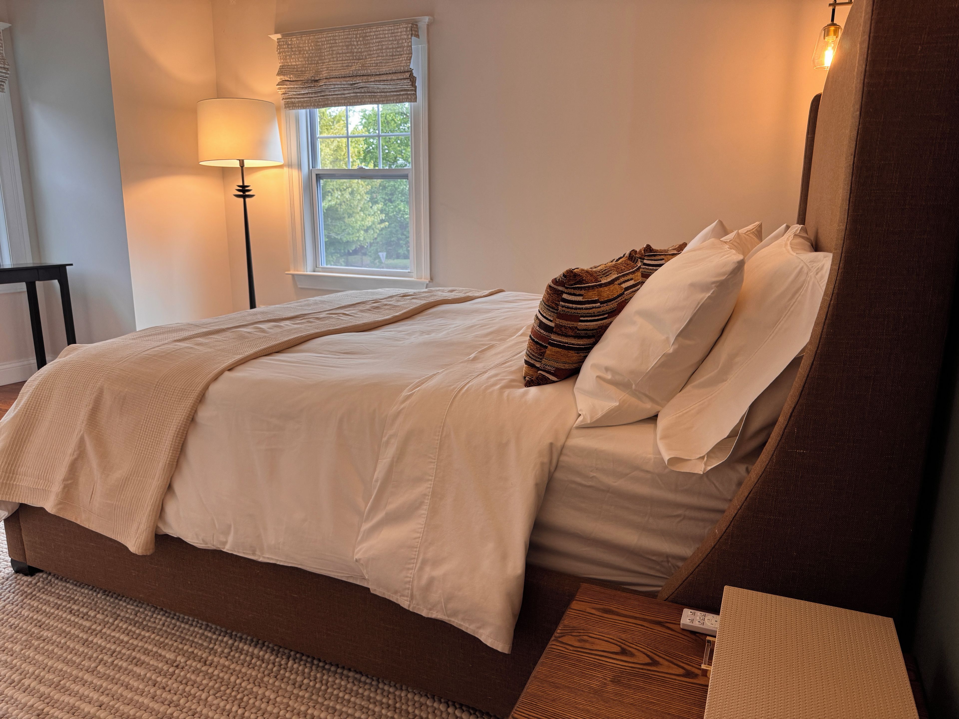 A bedroom with a large bed, a bedside table, a lamp, and a window with a view of trees.