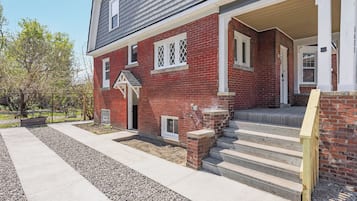 A two-story brick house with a front porch, a set of concrete steps leading to the entrance, and a small garden area with gravel and plants.