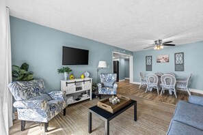 A living room with a flat-screen TV, a coffee table, patterned chairs, a dining area with white chairs, and a ceiling fan. - Emerald Escape at Sugar Beach C27 - Gulf Views, Beachfront Pool, Private Balcony (Panama City Beach)