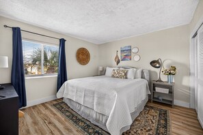 A bedroom with a bed, a nightstand, a lamp, a window with curtains, a wall hanging, and a rug. - Emerald Escape at Sugar Beach C27 - Gulf Views, Beachfront Pool, Private Balcony (Panama City Beach)
