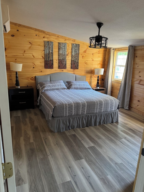 A bedroom with a wooden wall, a bed with a grey bedspread, two bedside tables with lamps, and a ceiling light fixture.