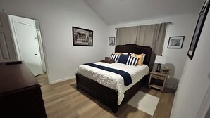 A bedroom with a bed, a nightstand, a lamp, and a door. - Furnished 2BR + Office/Guest Room Near Texas Health HEB & DFW Airport (Bedford)