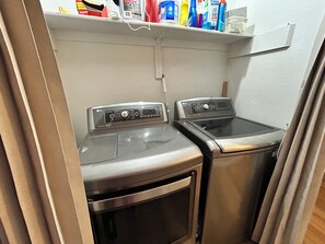 Un espace buanderie comprenant deux machines à laver et un sèche-linge, une étagère au-dessus pour ranger les produits de nettoyage, et un mur blanc.