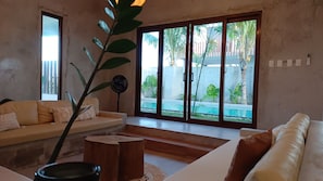 A living room with a sofa, a coffee table, and a fan. There is a large window with a view of a pool and palm trees.
