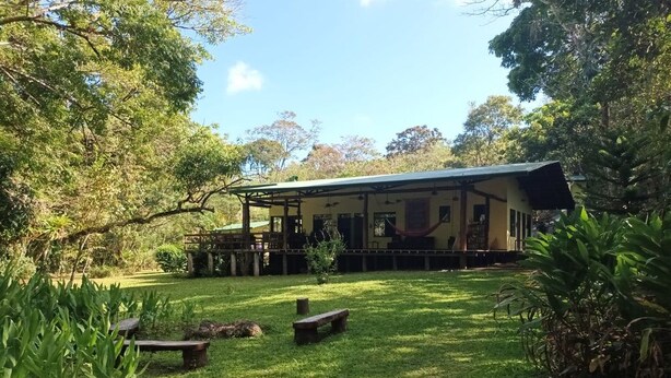 A building with a covered porch, surrounded by a grassy lawn and trees. - Rainforest Riverside Retreat Lake Arenal. Enjoy your family & friends getaway! (Tilaran)