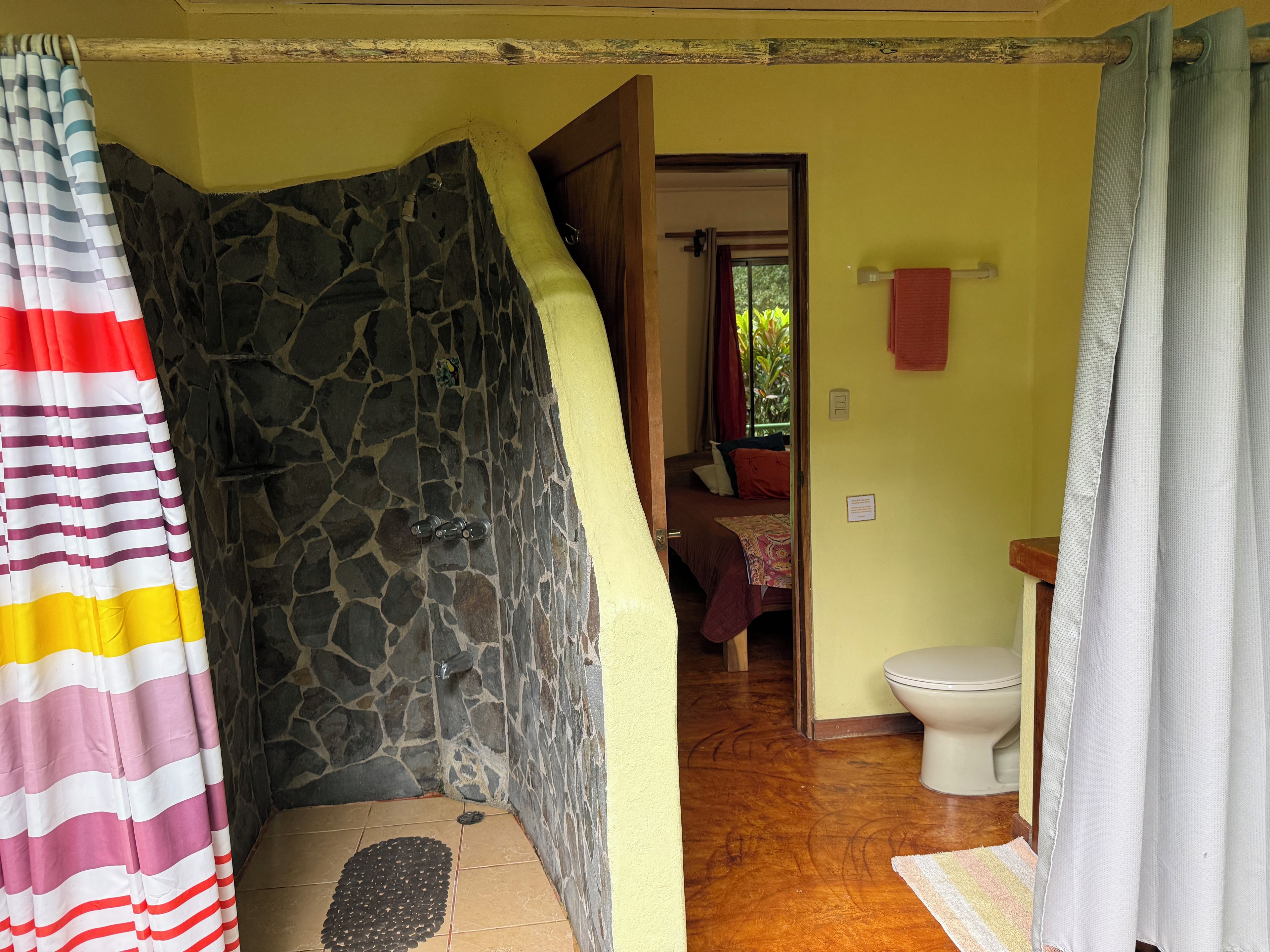 Une salle de bain avec un mur en pierre, un rideau de douche, des toilettes et un lit à l’arrière-plan.
