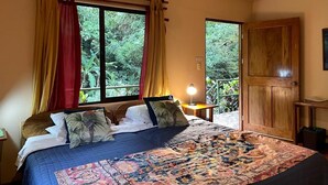 A bedroom with a large bed, curtains, a bedside table with a lamp, and a view of greenery through the window. - Rainforest Riverside Retreat Lake Arenal. Enjoy your family & friends getaway! (Tilaran)