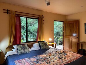 Ein Schlafzimmer mit einem großen Bett, Vorhängen, einem Nachttisch mit Lampe und Blick auf Grün durch das Fenster.