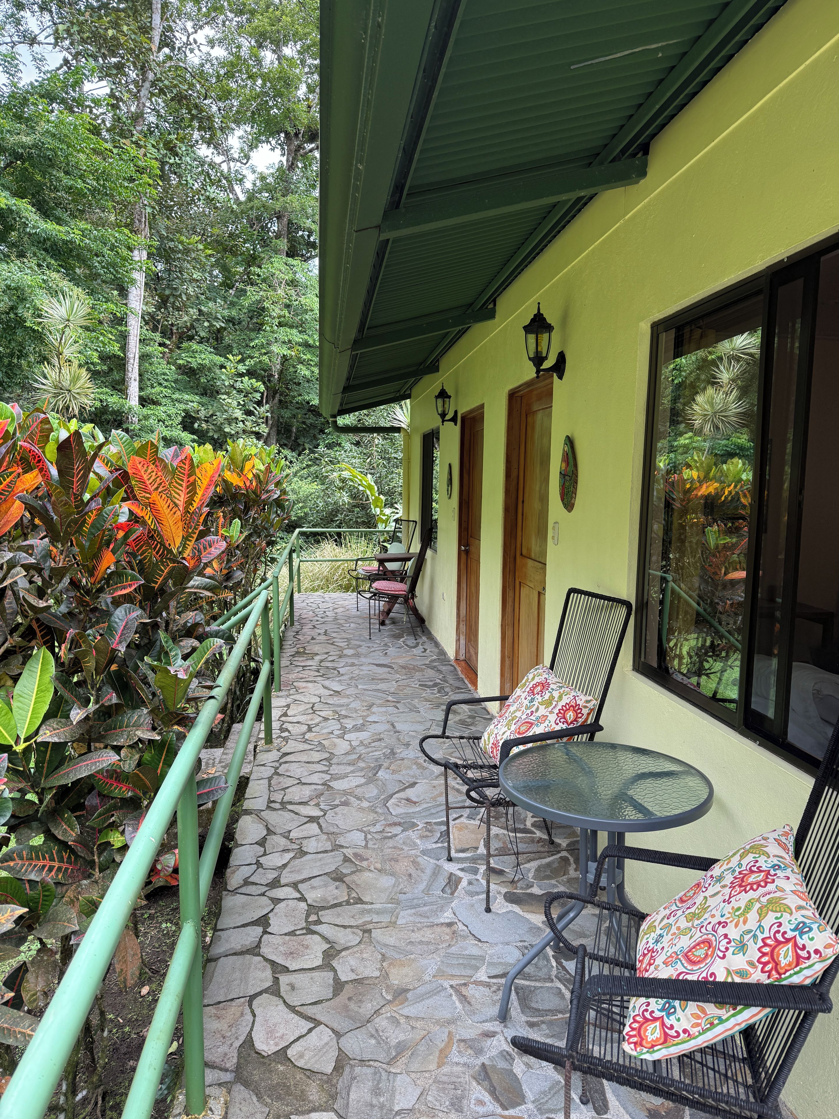 A covered outdoor area with stone flooring, two tables with chairs, and a variety of tropical plants.