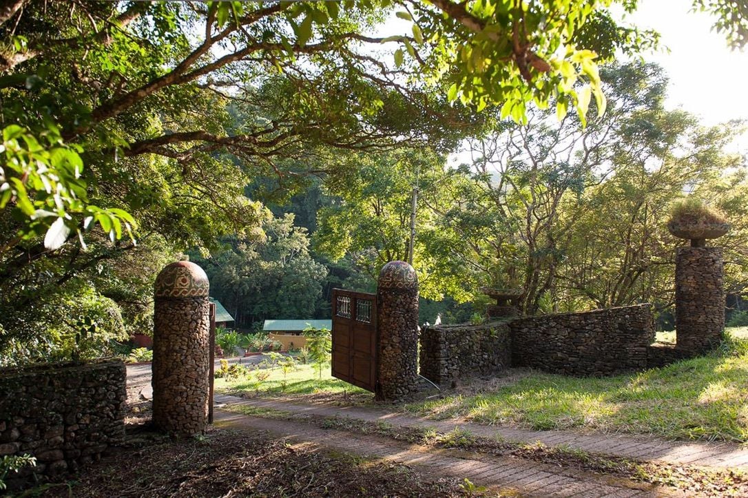 Une porte en pierre au design traditionnel, qui mène à un sentier bordé d’une végétation luxuriante et d’arbres.