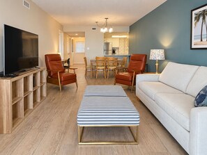 Ein Wohnzimmer mit Fernseher, zwei Ledersesseln, einem gestreiften Hocker, einem weißen Sofa und einem Essbereich mit Tisch und Stühlen.