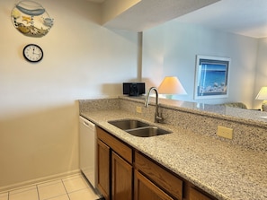 A kitchen with granite countertops, a stainless steel sink, wooden cabinets, a dishwasher, a wall clock, a small television, and a framed picture. - Land's End Resort - King Suite w/ Peaceful Views | Perfect for Couples (Myrtle Beach)
