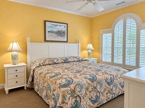A bedroom with a bed, two nightstands, a ceiling fan, a window with shutters, and a framed picture on the wall. - Land's End Resort - Secluded 2BR Condo | Relaxing Getaway (Myrtle Beach)