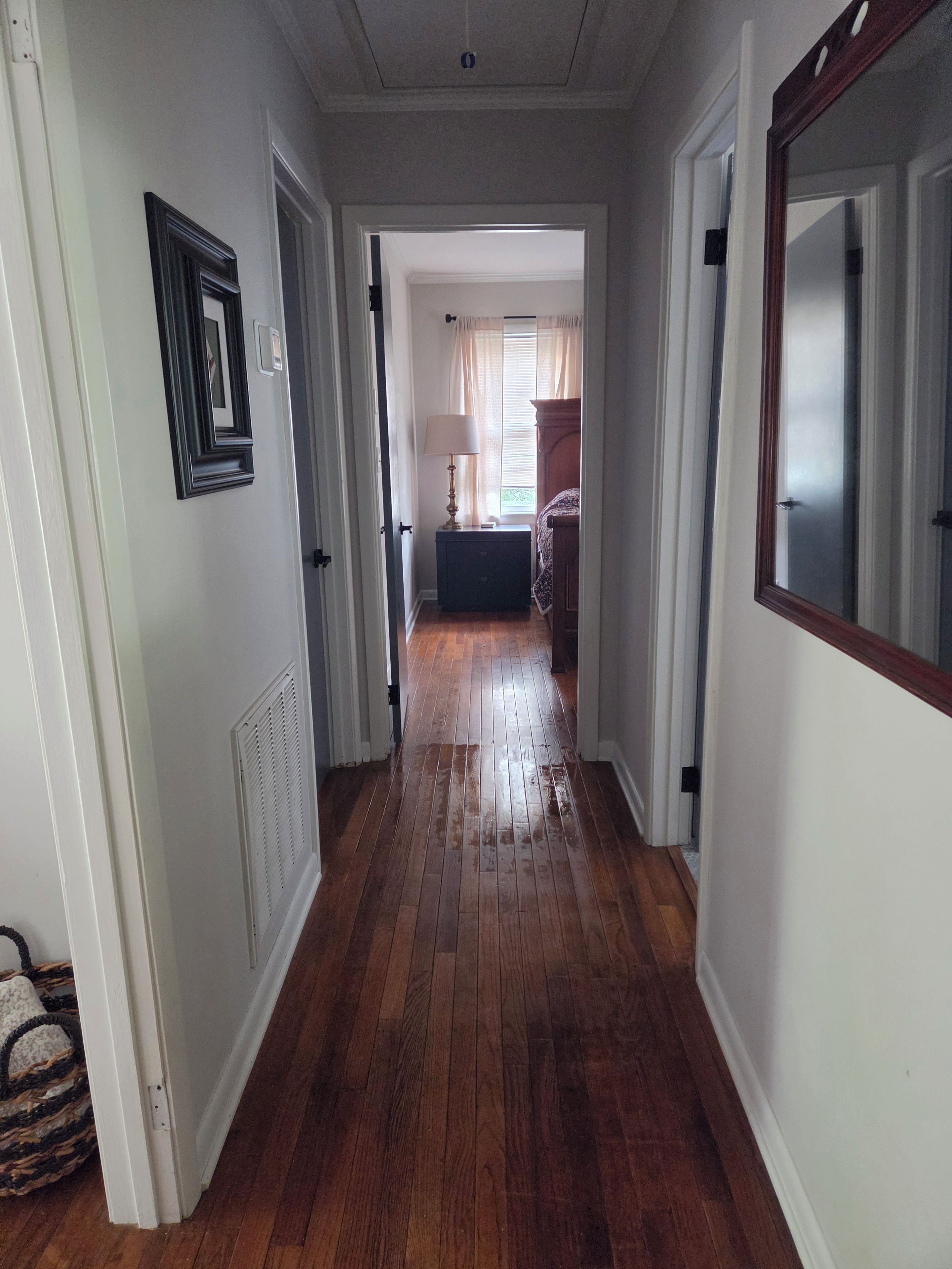 A hallway with wooden flooring, white walls, and a door leading to a bedroom with a bed and a nightstand.