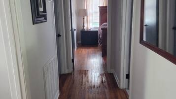 A hallway with wooden flooring, white walls, and a door leading to a bedroom with a bed and a nightstand.