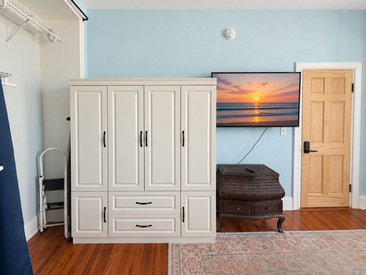 Une pièce avec une armoire blanche, une commode en bois, un téléviseur fixé au mur et une porte.