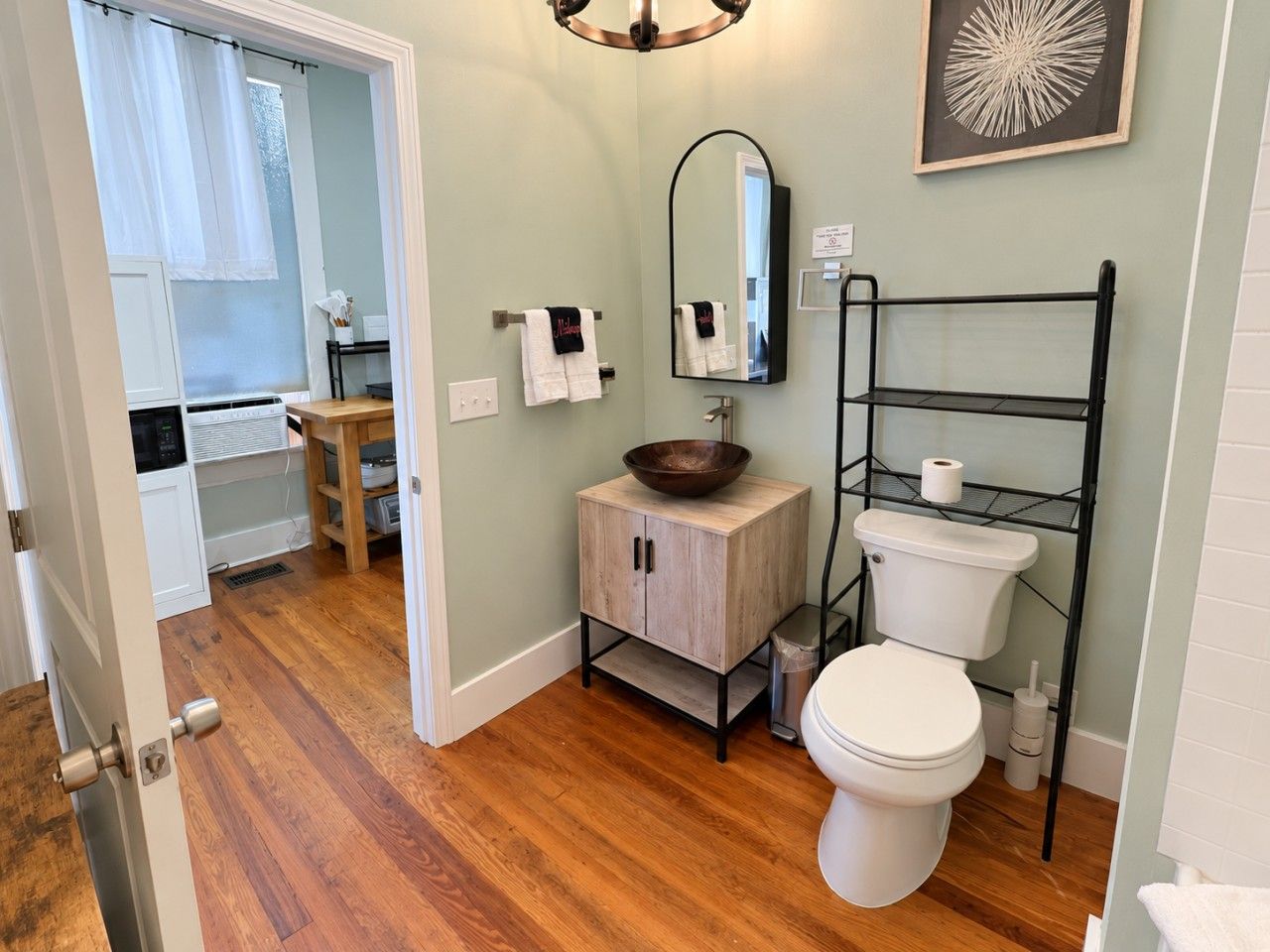 Une salle de bain comprenant des toilettes, un lavabo avec un meuble en bois, un miroir et une étagère en métal noir où est posé du papier toilette.
