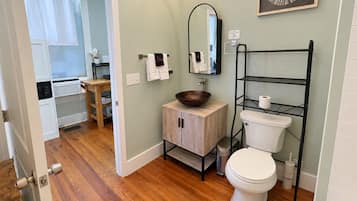 Une salle de bain comprenant des toilettes, un lavabo avec un meuble en bois, un miroir et une étagère en métal noir où est posé du papier toilette.