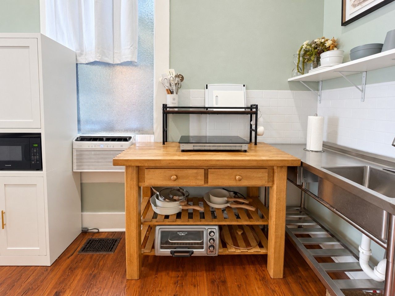 Une cuisine avec une table en bois, des placards blancs, un four à micro-ondes, un climatiseur et un évier en acier inoxydable.