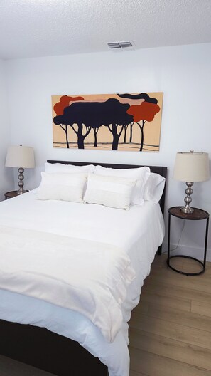 A neatly made bed with white linens, two bedside lamps, and a wall-mounted painting of trees. - "BeeNBee" 3-Bedroom Duplex In the heart of Ybor. (Tampa)