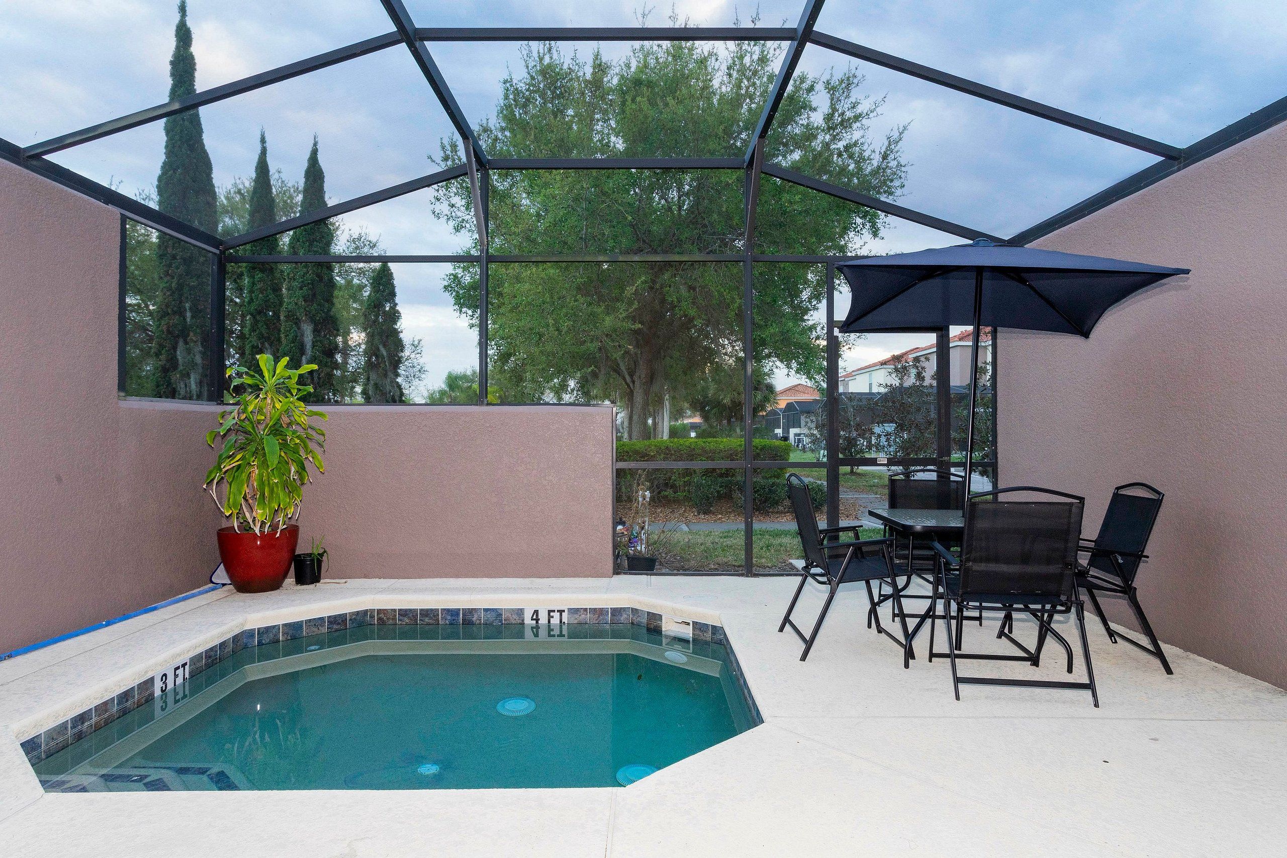 A glass-enclosed swimming pool with a hot tub, surrounded by a patio with chairs and an umbrella.