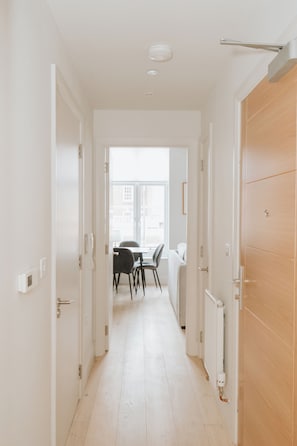 A hallway with white walls, a wooden door on the right, and a window at the end leading to a dining area with chairs and a table. - Experience London in Luxury – 1-Bed Apt (London)