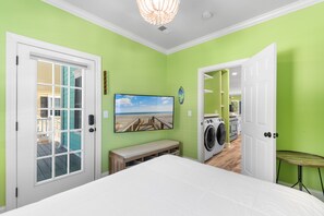 A room with a bed, a bench, a washing machine, and a door leading to another room. - Private Pool and Hot Tub — 5BR/4BA Beach House with Ocean Views in Surfside Beac (Surfside Beach)
