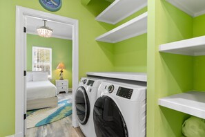 A laundry room with two front-loading washing machines, a bed with white bedding, a nightstand with a lamp, and a window with blinds. - Private Pool and Hot Tub — 5BR/4BA Beach House with Ocean Views in Surfside Beac (Surfside Beach)