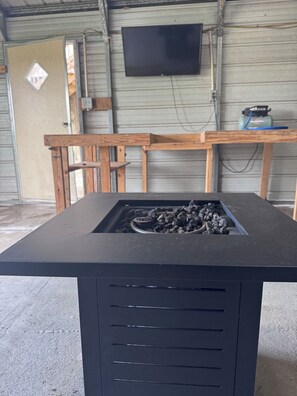 A black metal structure with a grill and charcoal, a wooden bench, and a television mounted on a metal wall.