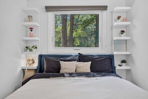Un dormitorio con cama, ventana con vista a árboles, estanterías blancas y cabecera azul.