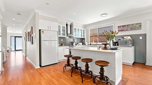 A modern kitchen with a central island, bar stools, and a large window. - The Sanctuary House By Book Jervis Bay (Sanctuary Point)