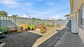 A backyard with a patio, a wooden bench, potted plants, and a metal fence. - The Sanctuary House By Book Jervis Bay (Sanctuary Point)