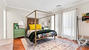 A four-poster bed with a floral bedspread, a green nightstand with a lamp, a wooden floor, and a large window with white curtains. - The Sanctuary House By Book Jervis Bay (Sanctuary Point)