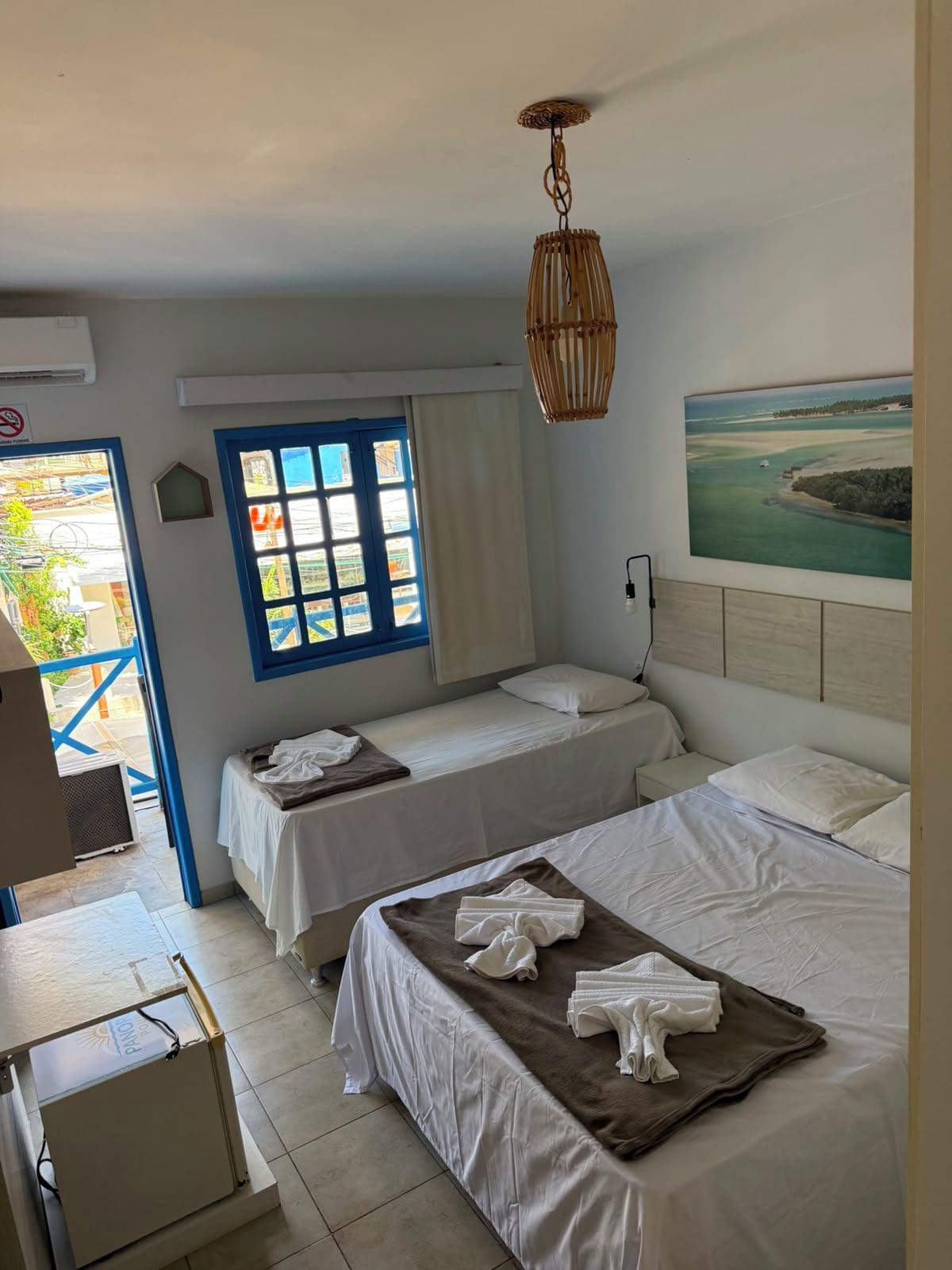 A hotel room with two beds, a painting of a coastal landscape, a window with blue shutters, and a ceiling fan.