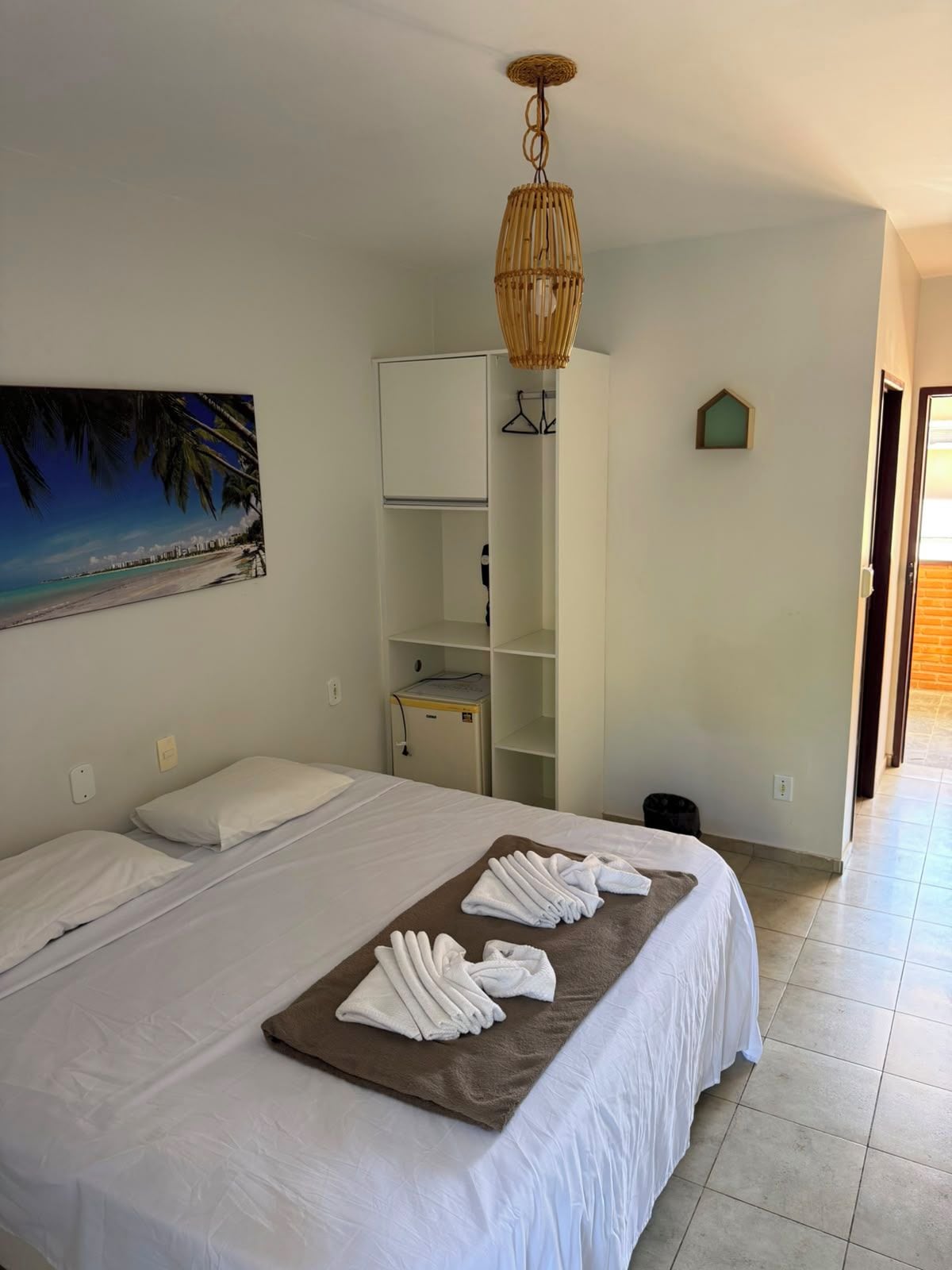 Quarto de hotel com cama, quadro de uma praia, guarda-roupa e luminária pendente.