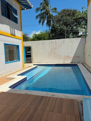 Una piscina con borde de azulejos, una terraza de madera y un edificio blanco con ventanas de marco azul.