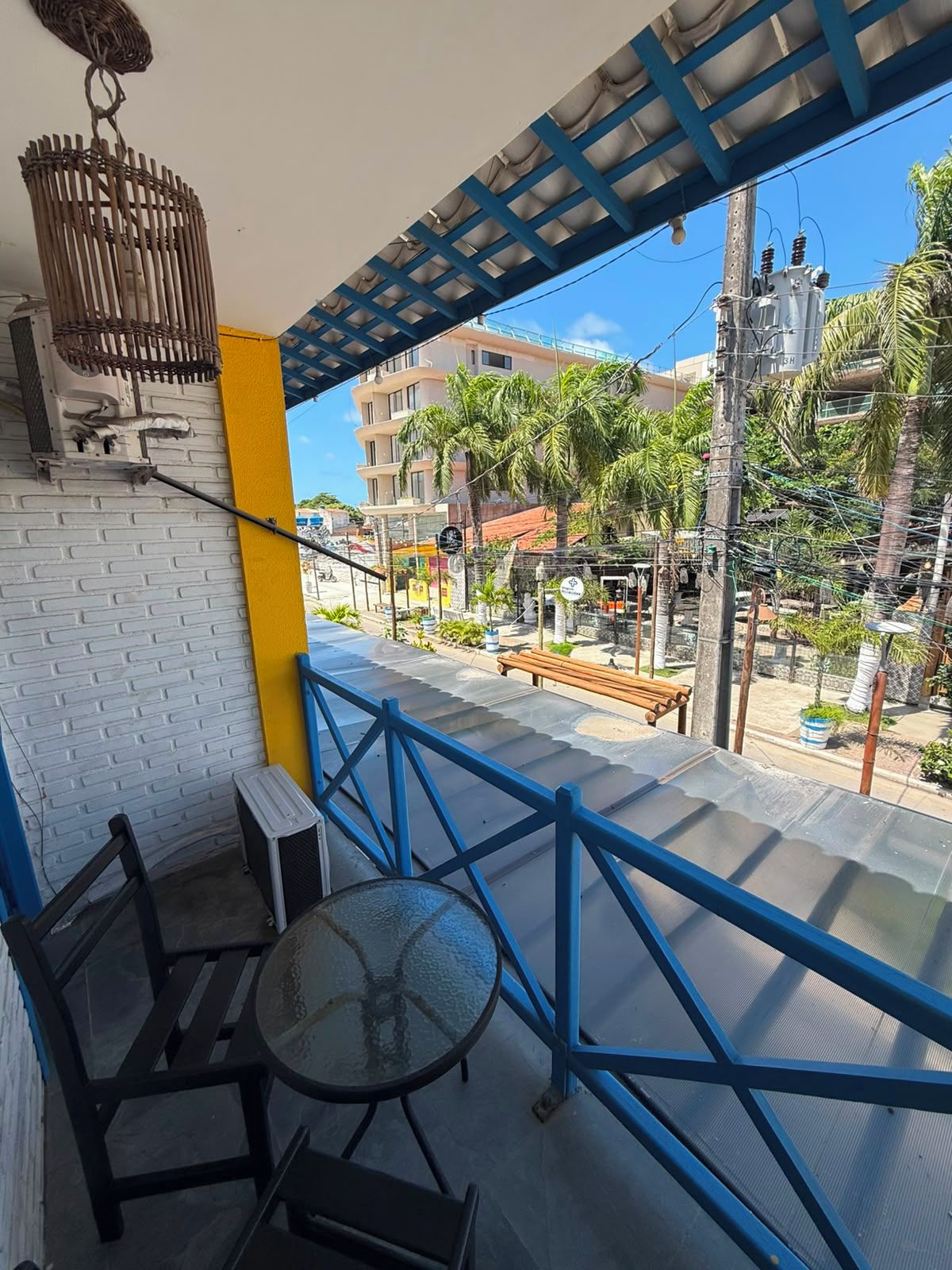 Varanda com mesa e cadeiras, com vista para uma rua com prédios e palmeiras.